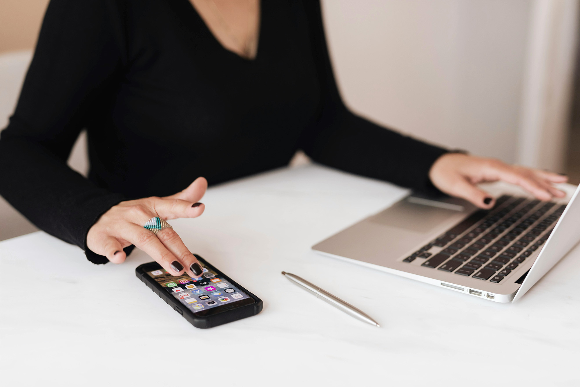 Woman wearing a black sweater, only the hands are shown. One hand is on the keyboard of her latpop, the other one is on the phone she has and she is scrolling something.