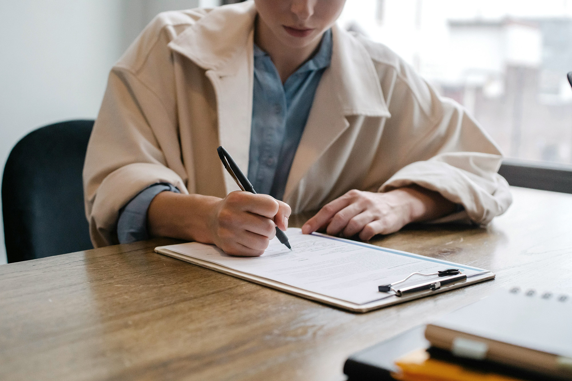 A woman is filling a form, she using a white coat and blue shirt and holding a pen while she is seating on a black chair. The form is on a desk made of wood.